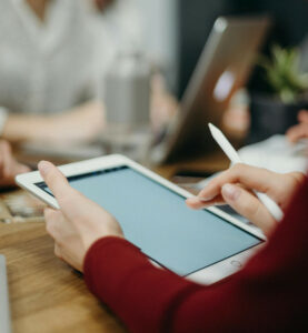 Close-up from person holding a tablet and pen. More people working in the background are blurred.